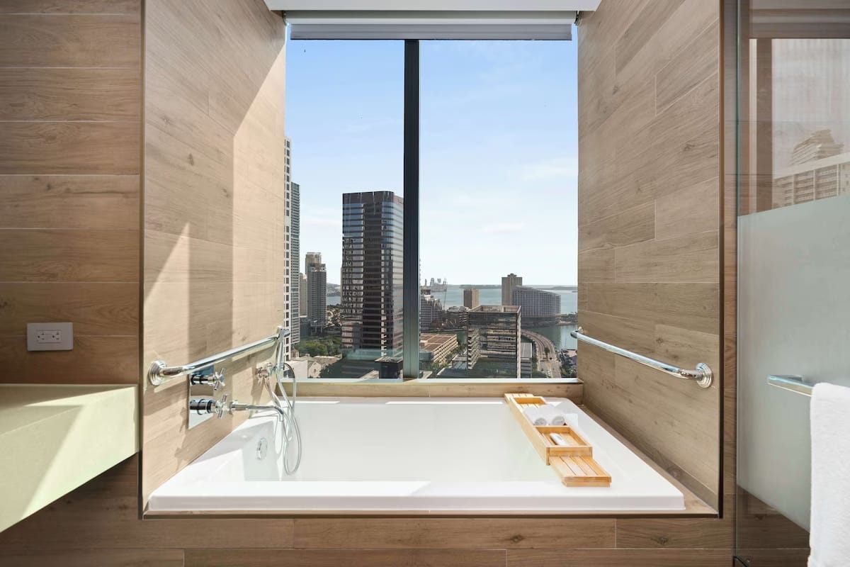 Luxury bathroom at EAST Miami in Brickell with soaking tub, floor-to-ceiling window, and skyline and bay views over downtown Miami.